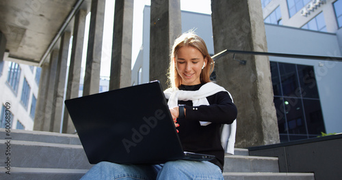 Portrait of good-looking calm smiling 20-aged light-haired girl in casual clothes which sitting on building's steps,working on laptop and checking time on handclock