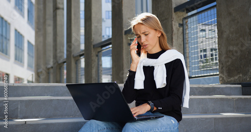 Portrait of adorable joyful smart modern 25-aged woman in trendy youthful wear which resting on concrete stairs of city building,revisioning information on laptop during mobile conversation