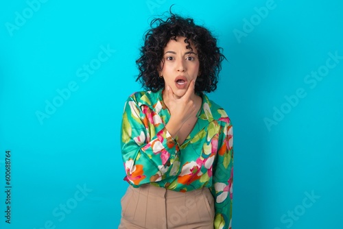 young arab woman wearing colorful shirt over blue background Looking fascinated with disbelief, surprise and amazed expression with hands on chin