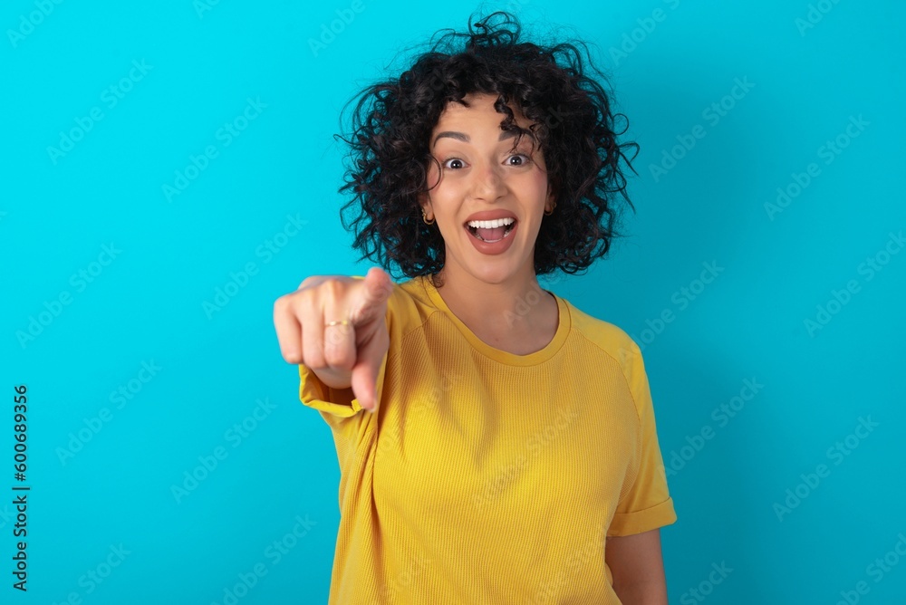 Excited positive young arab woman wearing yellow T-shirt over blue background points index finger directly at you, sees something very funny. Wow, amazing Excited positive young arab woman wearing yellow T-shirt over blue background points index finger directly at you, sees something very funny. Wow, amazing