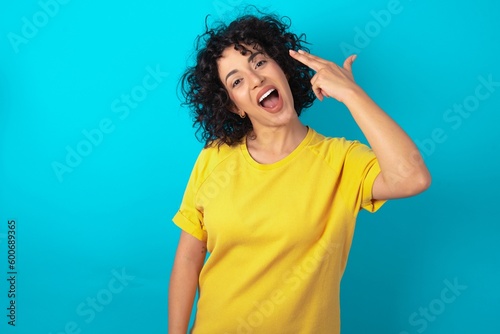 Tablou pe pânză young arab woman wearing yellow T-shirt over blue background foolishness around shoots in temple with fingers makes suicide gesture