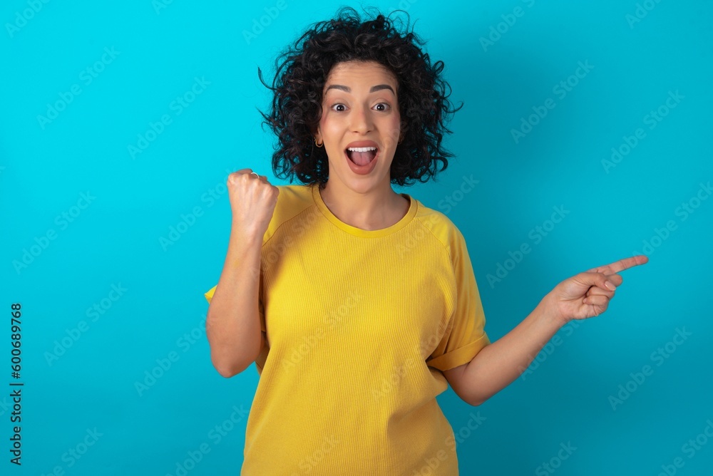 young arab woman wearing yellow T-shirt over blue background points at empty space holding fist up, winner gesture. young arab woman wearing yellow T-shirt over blue background points at empty space holding fist up, winner gesture.
