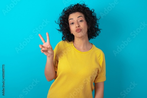 young arab woman wearing yellow T-shirt over blue background makes peace gesture keeps lips folded shows v sign. Body language concept