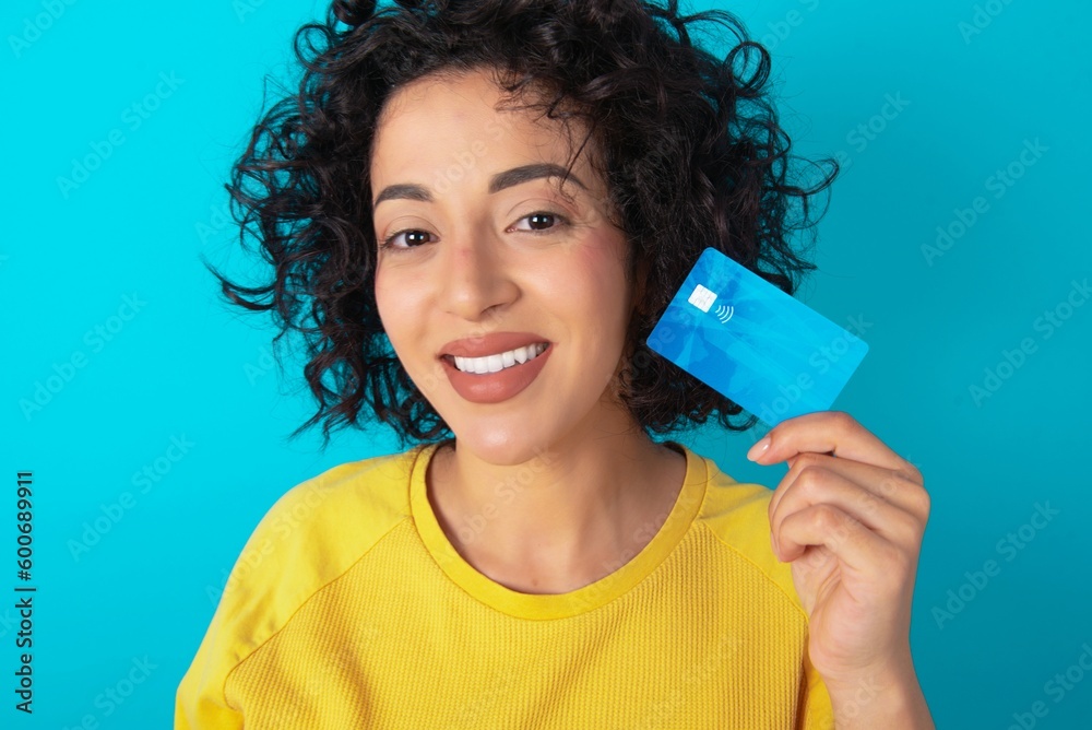 Close up photo of optimistic young arab woman wearing yellow T-shirt over blue background hold card Close up photo of optimistic young arab woman wearing yellow T-shirt over blue background hold card