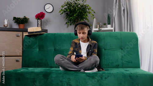 Good-looking stylish concentrated 10-12-aged boy in earphones relaxing on comfortable sofa in beautifully designeted living-room and enjoying modern songs from smartphone