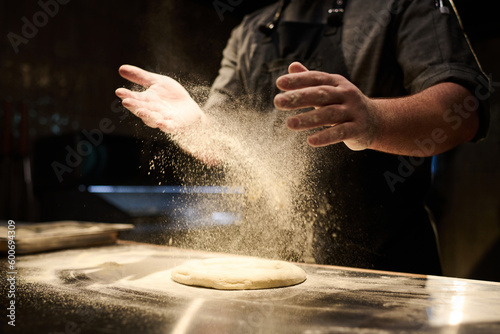 hands of young male chef sprinkling flour on dough flatbread before kneading it on table in the kitchen while preparing pizza