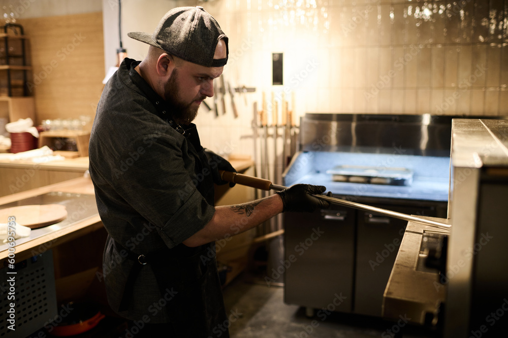 Side view of young cook using metallic holder while standing in the kitchen of restaurant and taking baked pizza out of hot electric oven Side view of young cook using metallic holder while standing in the kitchen of restaurant and taking baked pizza out of hot electric oven