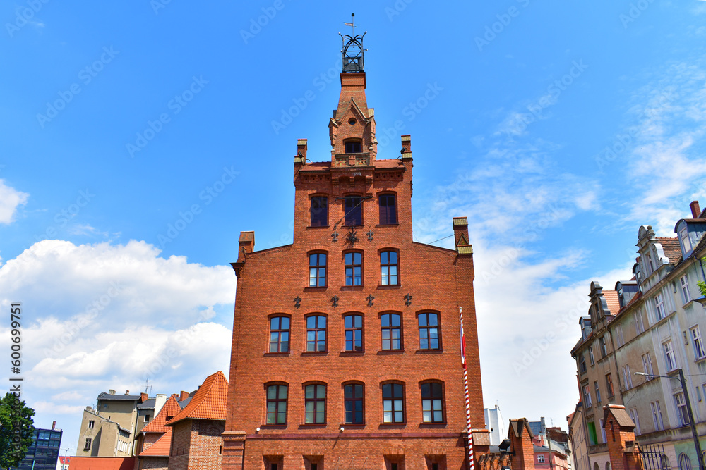 Obraz premium Old architectural building made of red bricks, windows and a tower at the top with forged elements. Sunny summer day with blue sky. Poland, Poznan, June 2022