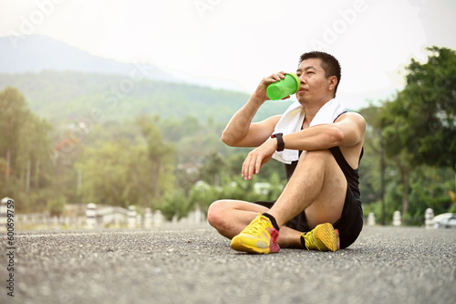 Sweaty and thirsty mature Asian man sits on the road in a public park and drinks water