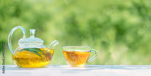 glass teapot and cup with herbal tea close up on table, natural abstract green background. summer season. relax time. useful calming tea. Tea party in garden. template for design. copy space