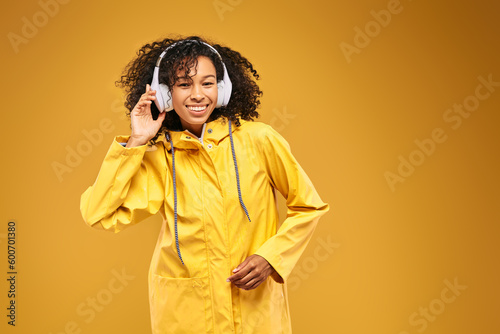 Beautiful African woman in yellow raincoat listening to music in headphones while standing in studio on yellow background