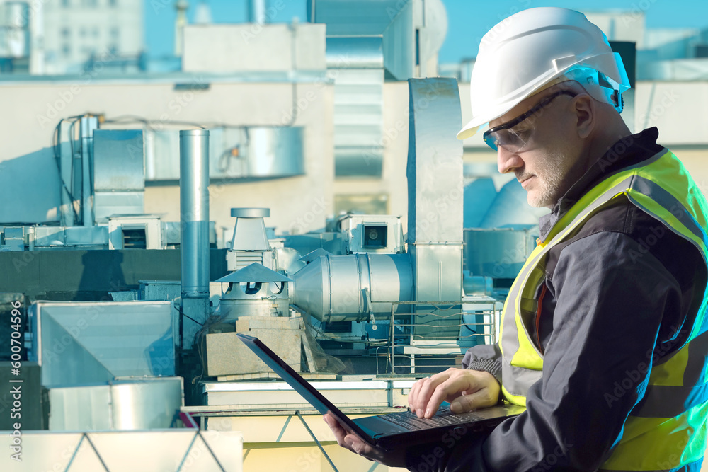 Engineer with laptop. Man on roof of building. Specialist oversees operation ventilation. Master in installation ventilation ducts. Ventilation engineer. Man in protective helmet on roof of factory
