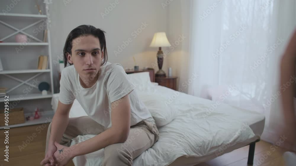 Careless young man sitting in bedroom as angry woman hitting with pillow crying leaving. Portrait of negligent Caucasian boyfriend arguing with girlfriend breaking up Careless young man sitting in bedroom as angry woman hitting with pillow crying leaving. Portrait of negligent Caucasian boyfriend arguing with girlfriend breaking up