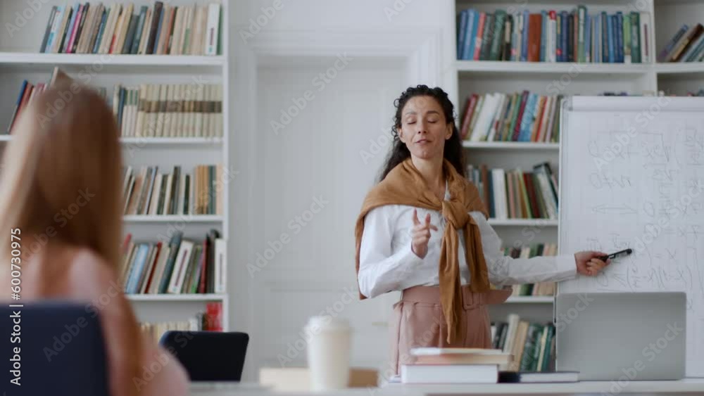 Professional young woman teacher explaining topic for students standing near whiteboard at classroom, girl raising hand Professional young woman teacher explaining topic for students standing near whiteboard at classroom, girl raising hand