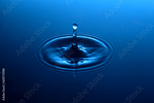A brilliant rich blue pool of water gently rippled with concentric circles with a single drop of connected spherical water splash above a column of liquid.