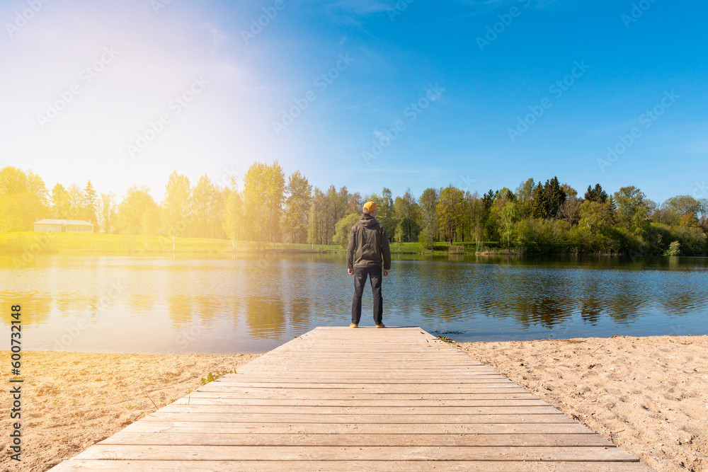 Young man standing alone on edge of wooden footbridge and staring at lake