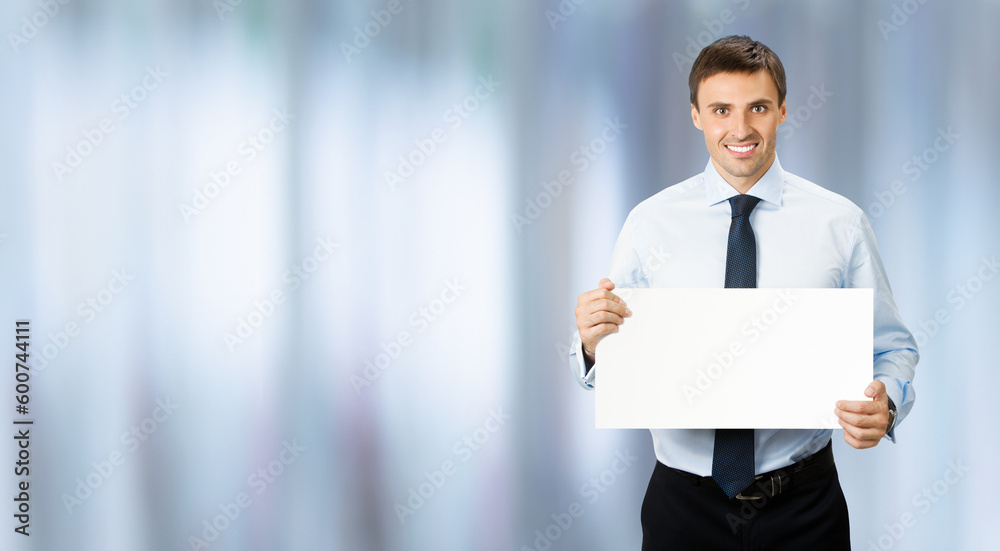 Image of business man professional bank manager in confident cloth, wear shirt, necktie tie. Standing businessman hold, show empty white banner sign board. Blurred office background. Image of business man professional bank manager in confident cloth, wear shirt, necktie tie. Standing businessman hold, show empty white banner sign board. Blurred office background.