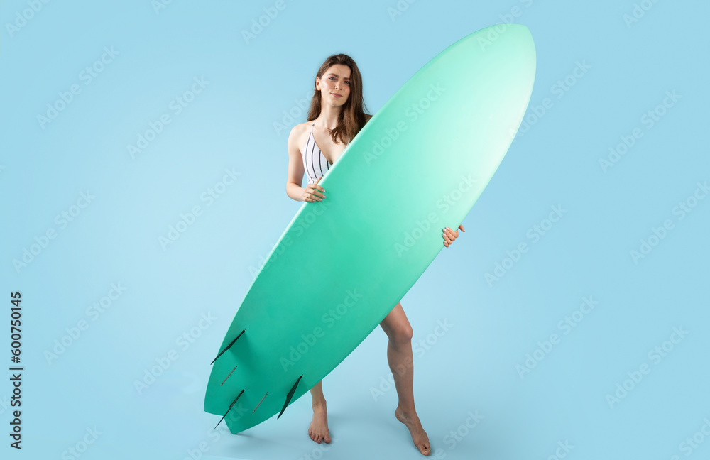 Summer extreme sport. Young lady in swimsuit posing with surfing board, enjoying summertime, pastel blue background