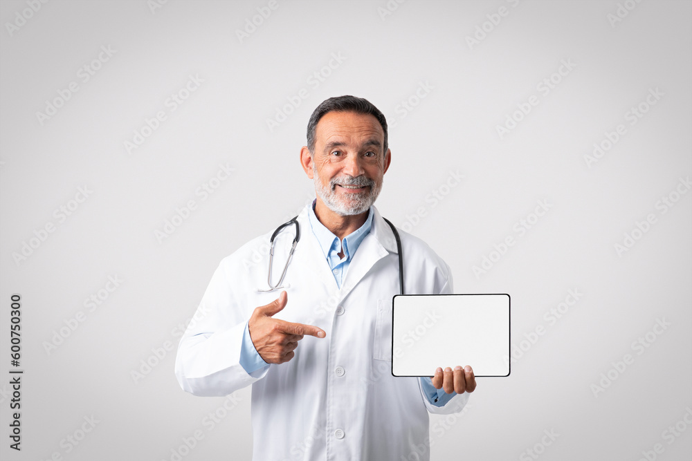 Cheerful european senior doctor in white coat with stethoscope points finger at tablet with empty screen Cheerful european senior doctor in white coat with stethoscope points finger at tablet with empty screen