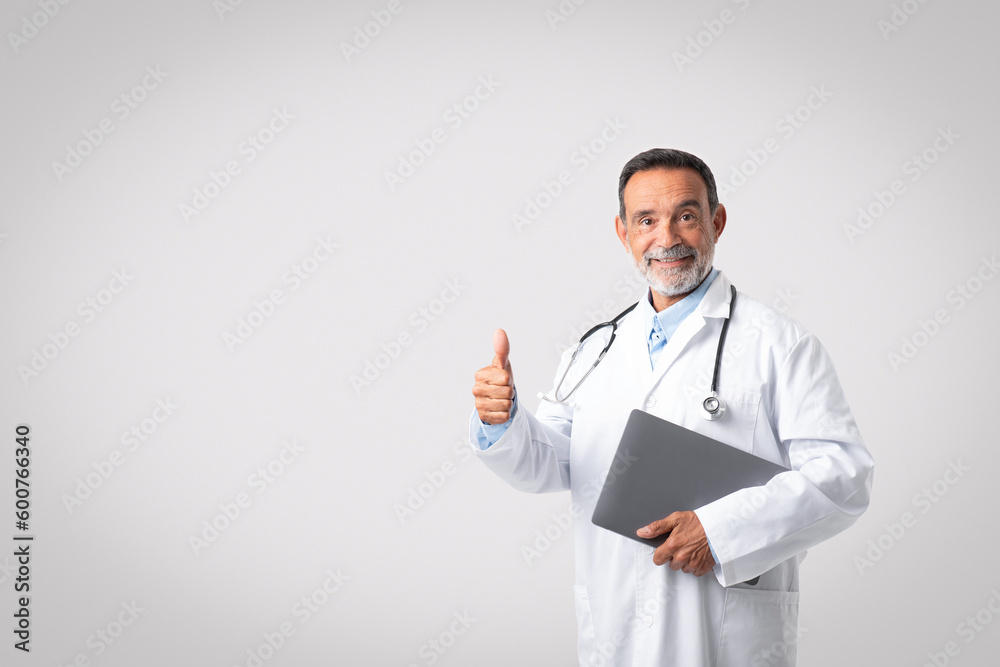 Smiling confident european senior man doctor in white coat with stethoscope and laptop show thumb up Smiling confident european senior man doctor in white coat with stethoscope and laptop show thumb up