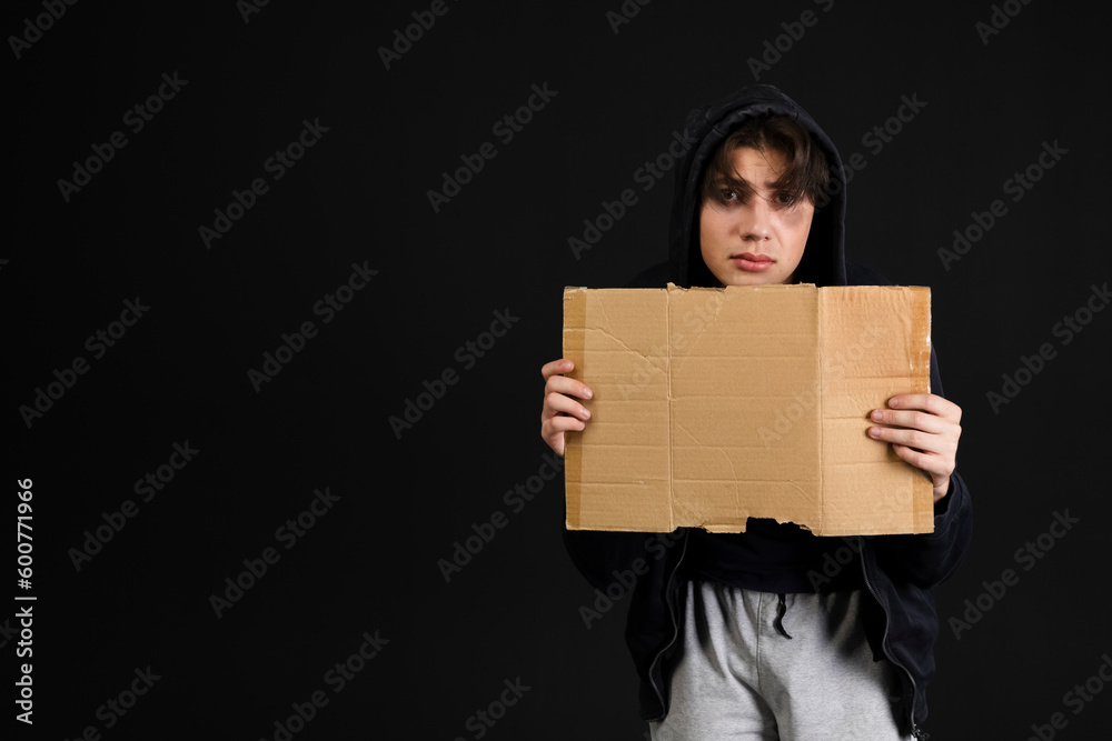A homeless young person with bruises under their eyes in dirty, tattered clothing, holding out an empty blank cardboard sign. Isolated on black background. A homeless young person with bruises under their eyes in dirty, tattered clothing, holding out an empty blank cardboard sign. Isolated on black background.