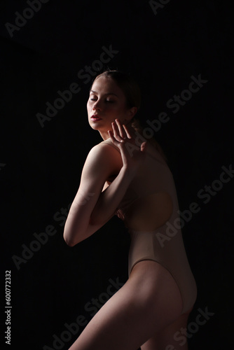 Ballerina in a beige bodysuit. Dark background. Sculpted beautiful female body. Pose of a gymnast.