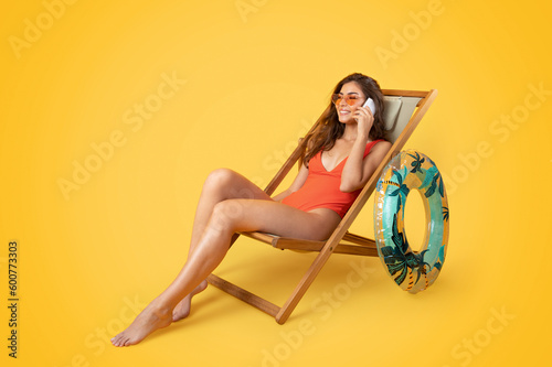 Positive millennial european woman in swimsuit, sunglasses with inflatable ring sit on deck chair #600773303