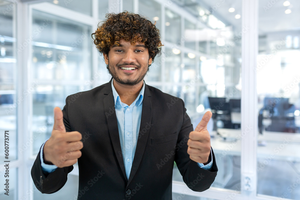 Fototapeta premium Portrait of successful hispanic businessman boss, manager in business suit and beard looking at camera and smiling standing near window, showing thumbs up, sign of success and achieving goals.