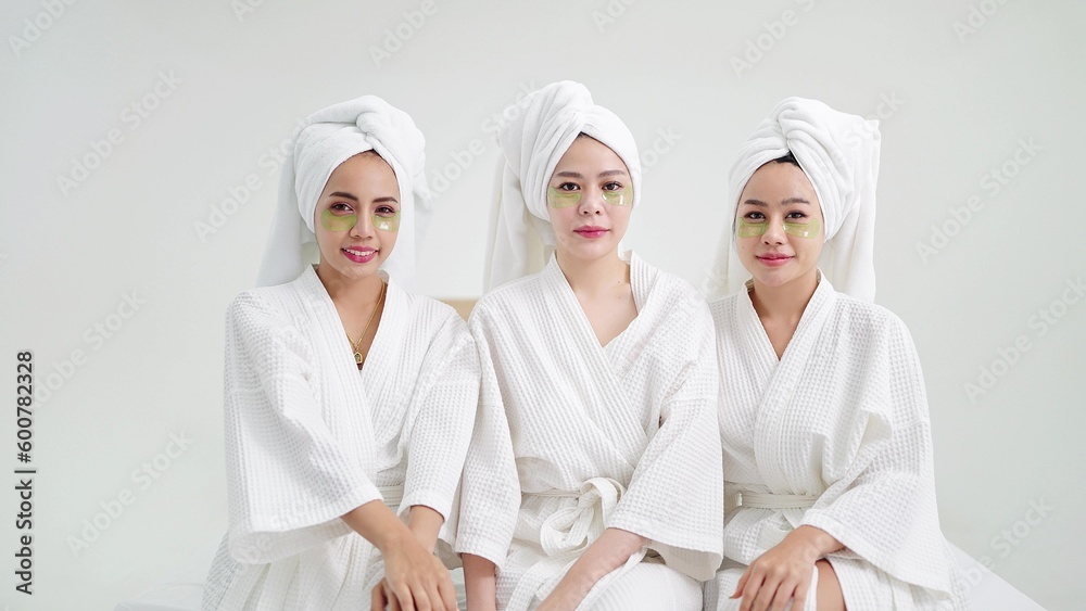 Group of beautiful young asian women in bathrobe and towels on heads with collagen patch on under eyes sitting and looking to camera. Self care and daily beauty routine