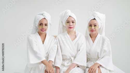 Group of beautiful young asian women in bathrobe and towels on heads with collagen patch on under eyes sitting and looking to camera. Self care and daily beauty routine