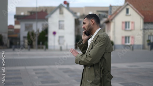 A Moroccan guy discussing on phone standing outside in city street. One Middle Eastern male person talking on smartphone device. Tracking shot in motion