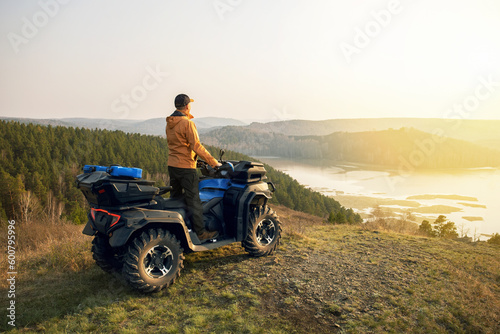 Man sitting on a quad bike at the top of a mountain and looking towards the lake at sunset.