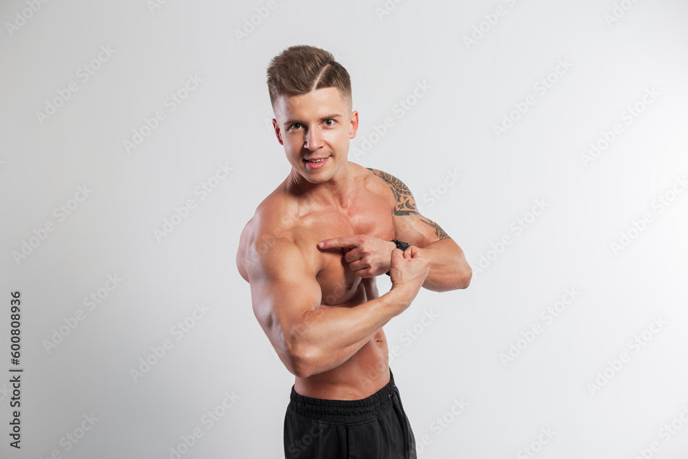 Fototapeta premium Handsome happy athletic muscular fit man with a beauty naked inflated torso shows the muscles of the biceps and points with a finger to the biceps on a white background in the studio. Workout time