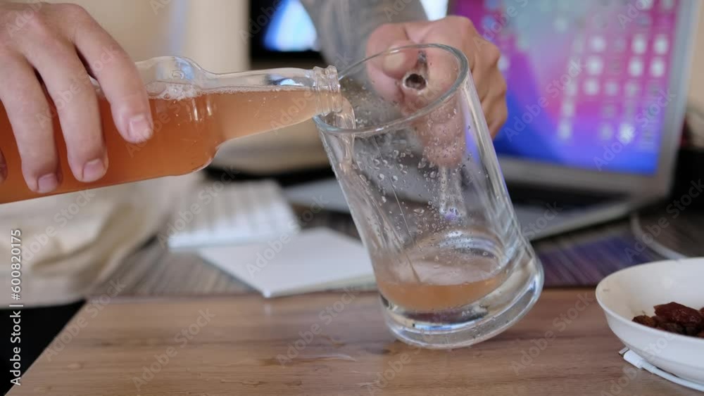 Man pouring cold craft beer from bottle into glass mug, against the background of a desktop with a lying laptop, motion close up. Process of filling cup with beer. Man pouring cold craft beer from bottle into glass mug, against the background of a desktop with a lying laptop, motion close up. Process of filling cup with beer.