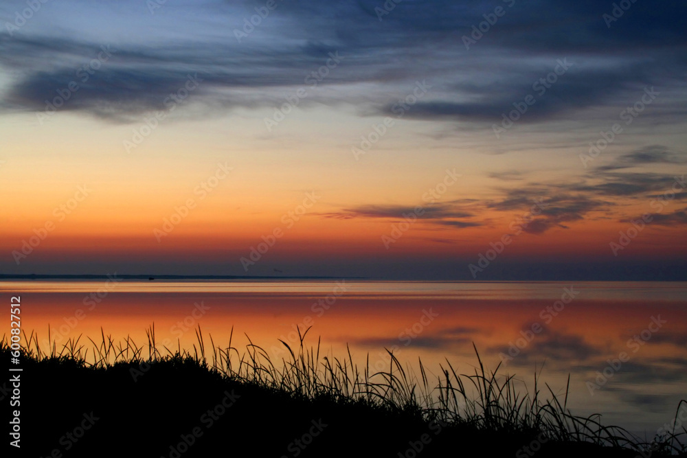 Early morning. The sun soon will rise above the sea and will illuminate clouds. In the quiet sea beautiful reflection. Pleasant colours. A quiet nature. Silhouettes of a grass on a coast