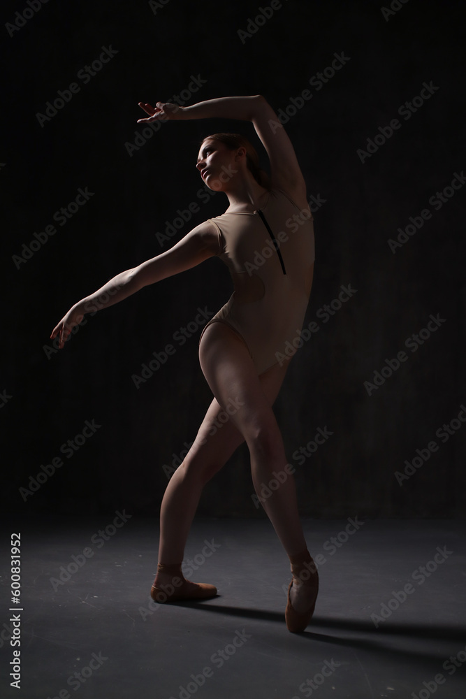Fototapeta premium Ballerina in a beige bodysuit and pointe shoes. Dark background. Sculpted beautiful female body. Pose of a gymnast.