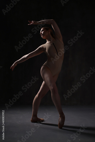 Fototapeta Naklejka Na Ścianę i Meble -  Ballerina in a beige bodysuit and pointe shoes. Dark background. Sculpted beautiful female body. Pose of a gymnast.