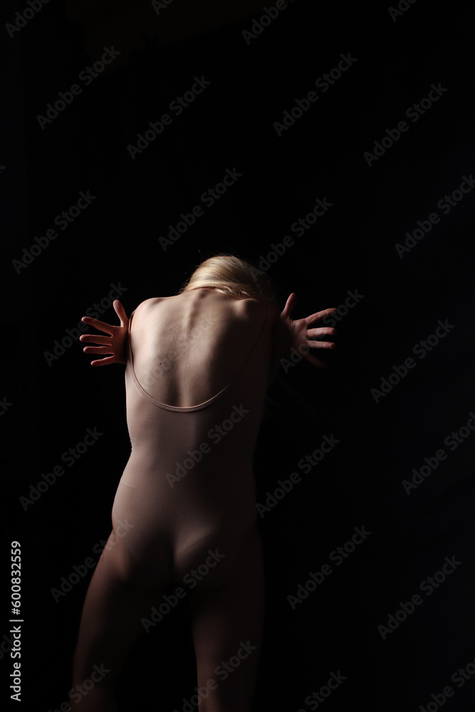 Ballerina in a beige bodysuit. Dark background. Sculpted beautiful female body. Pose of a gymnast. Ballerina in a beige bodysuit. Dark background. Sculpted beautiful female body. Pose of a gymnast.
