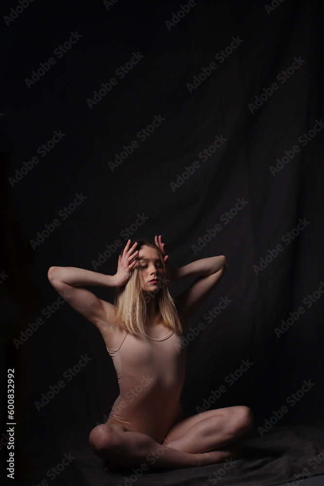 Ballerina in a beige bodysuit. Dark background. Sculpted beautiful female body. Pose of a gymnast.