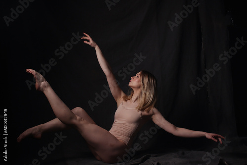 Ballerina in a beige bodysuit. Dark background. Sculpted beautiful female body. Pose of a gymnast.