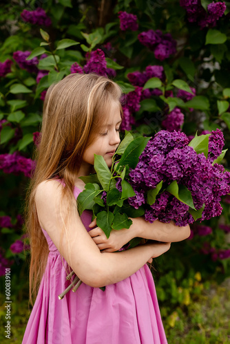 a beautiful happy smiling blonde girl with long hair in a pink dress with a purple bouquet of lilacs in her hands stands against the background of a lilac bush