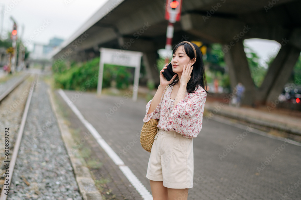 Asian young woman traveler with weaving basket using a mobile phone beside railway train station in Bangkok. Journey trip lifestyle, world travel explorer or Asia summer tourism concept.