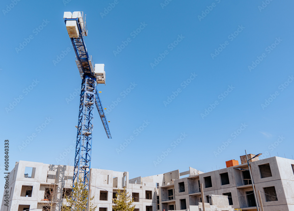 High-rise building crane with a long arrow of blue color on blue clear sky above a concrete building under construction with solid walls
