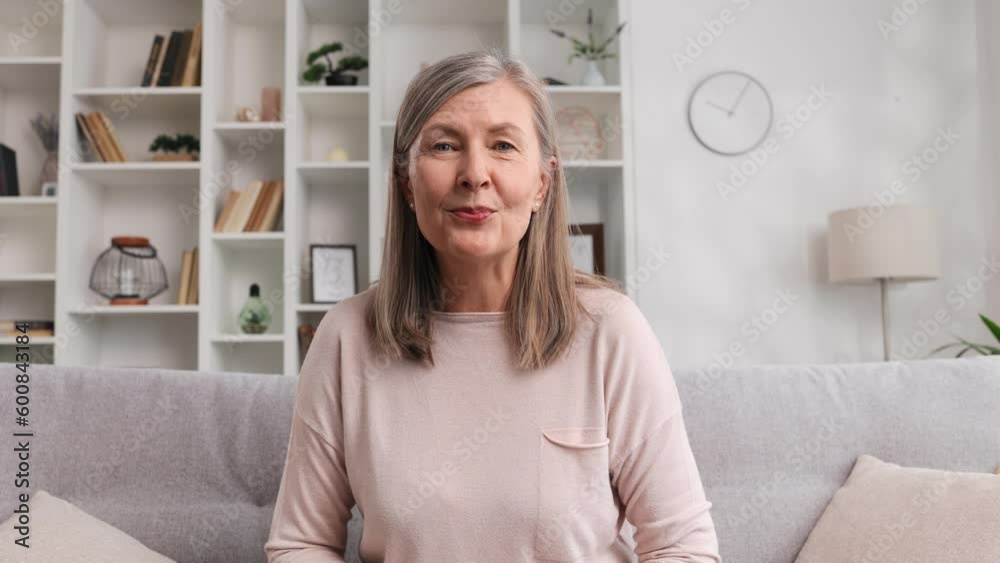 Joyful elderly woman in her 50s 60s looking at the camera at home during online call. Smiling senior grandmother talking to webcam, video chatting, chatting online, sitting on sofa, watching webcam.