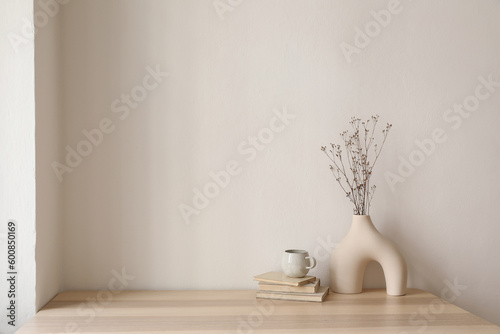 Elegant interior still life. Modern organic, geometric shaped vase with dry flowers, grass. Pile of old books, cup of coffee on wooden table. Home staging, minimal decor concept. Blank beige wall.