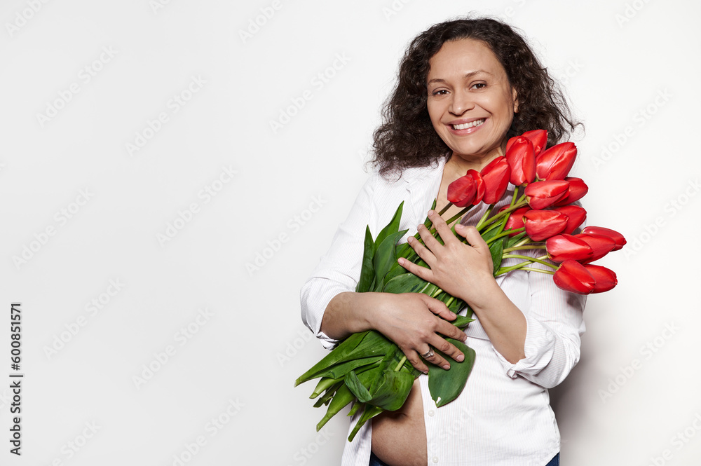 Happy delightful pregnant woman, wearing white unbuttoned shirt, holding a bunch of beautiful red tulips for special occasion, smiling isolated on white background. Happy Mother's Day. Copy ad space Happy delightful pregnant woman, wearing white unbuttoned shirt, holding a bunch of beautiful red tulips for special occasion, smiling isolated on white background. Happy Mother's Day. Copy ad space