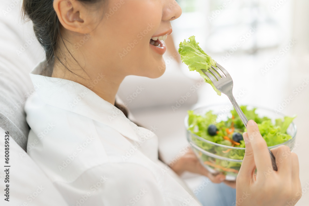Diet, Dieting asian young woman, girl hand use fork at lettuce on mix vegetables, green salad bowl, eat food low fat to good body sitting on sofa at home. Nutritionist female, Weight loss for healthy.