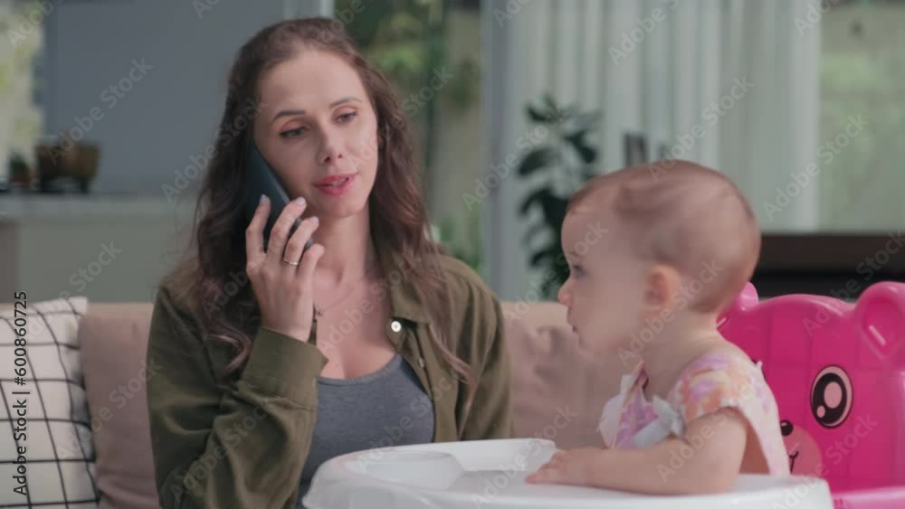 Young brunette woman with smartphone by ear talking to someone while sitting on couch in front of her cute baby daughter and looking at her during communication Young brunette woman with smartphone by ear talking to someone while sitting on couch in front of her cute baby daughter and looking at her during communication