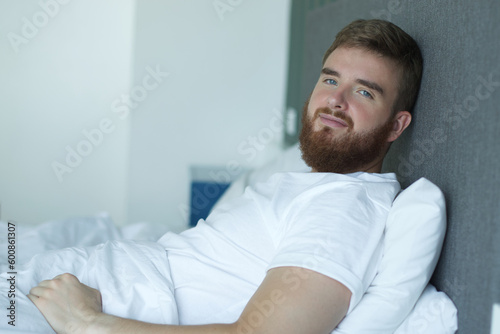 Young happy bearded handsome sleepy man lay in bed in bedroom at home in early morning, smile and looking at camera © Евгений Шемякин