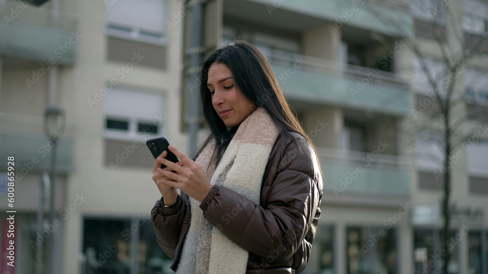 One young woman standing in city street looking at smartphone device. Person receiving positive notification on phone in urban setting